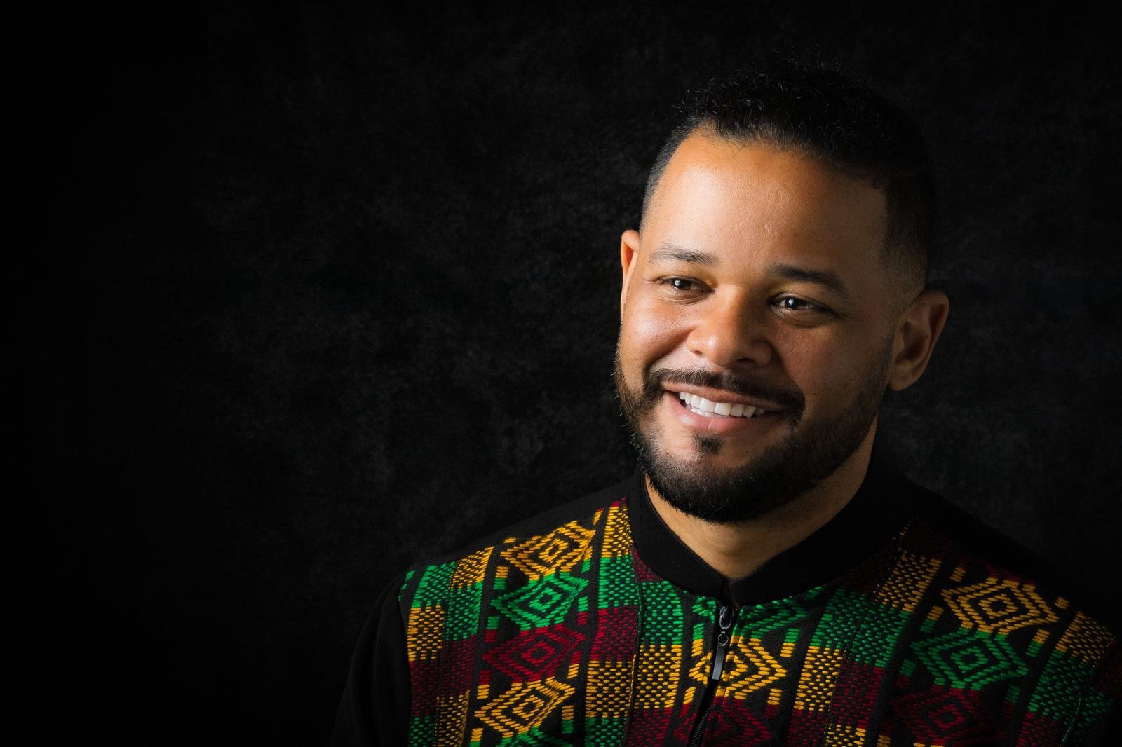 Matt smiles past the camera in a black jacket trimmed with green, gold and red woven pattern, side-lit against the dark studio backdrop.