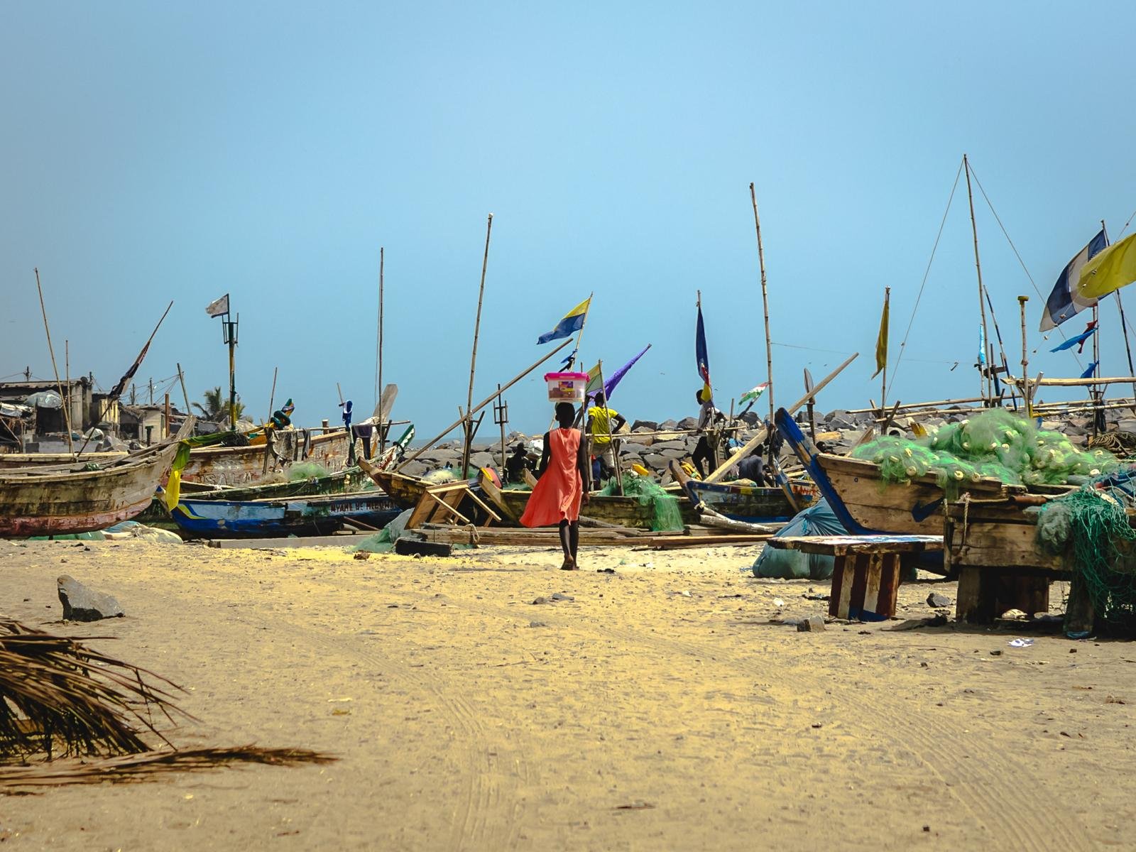 A woman in a coral dress carries a container on her head through a fishing harbour, wooden boats and flags behind her, nets piled in the sun.