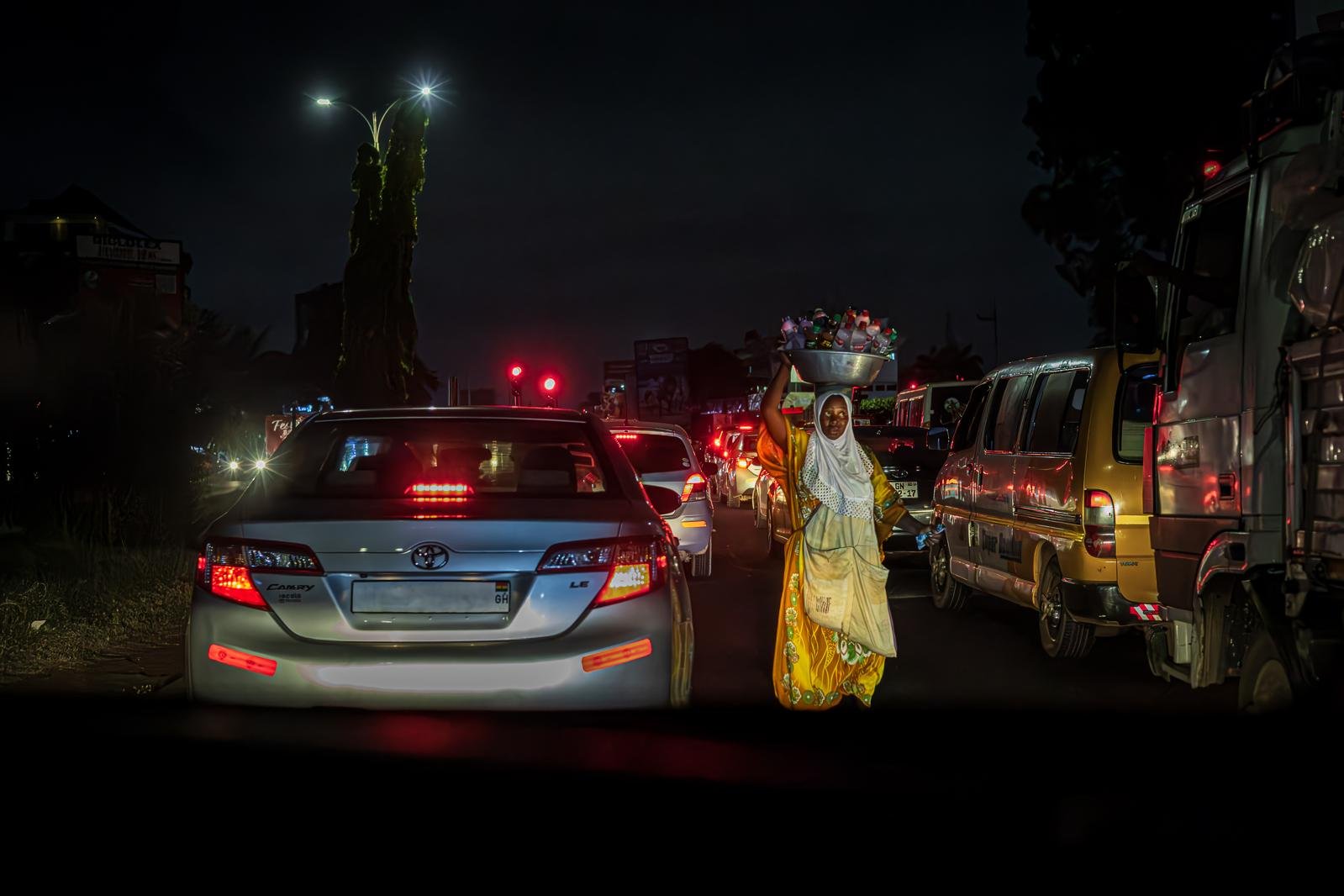In stopped night traffic, a woman in a white headscarf carries a basin of drinks on her head between the lanes, brake lights glowing around her.
