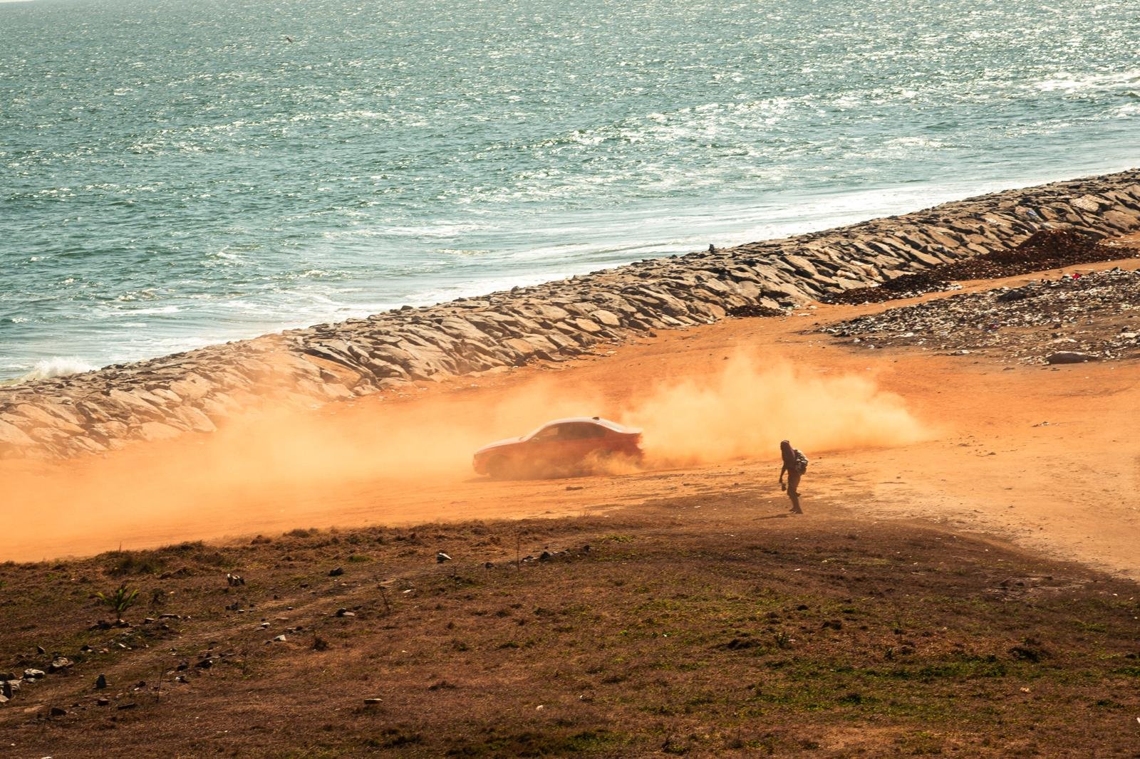 A red car trails a cloud of orange dust along the shore road as a lone figure walks through the haze, the sea bright beyond the breakwater.