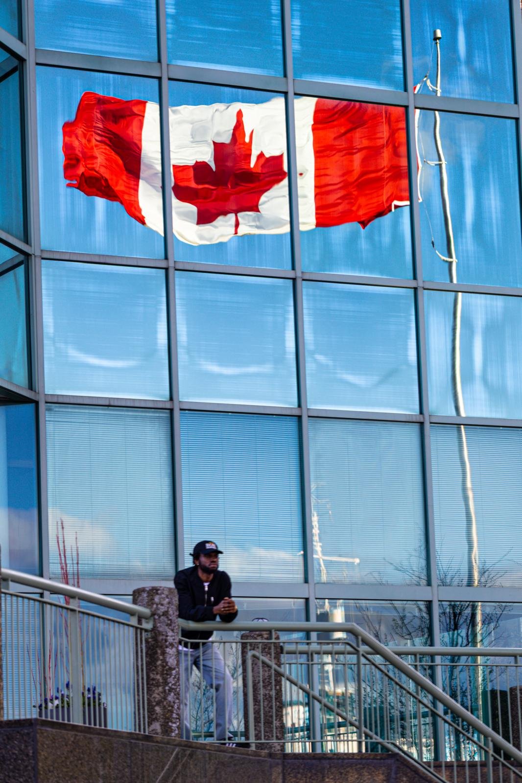 A man in a cap leans on a railing beneath a giant Canadian flag reflected in the glass of an office tower.