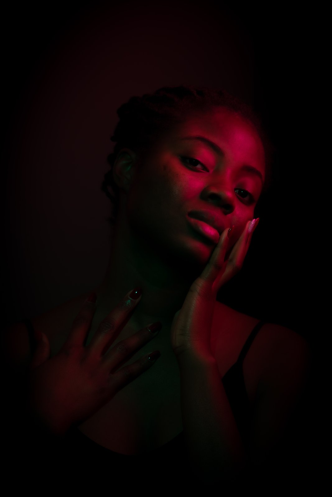 Portrait in red gel light with a green edge. She rests her head against one hand at her cheek, eyes calm.