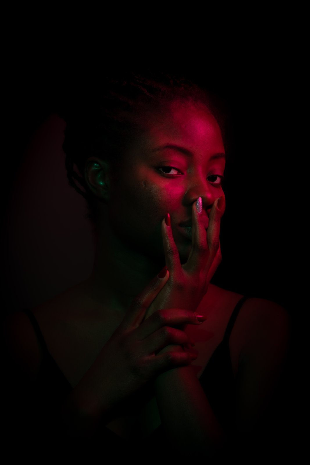 Close portrait in red gel light. Two fingers rest across her lips, one nail catching a silver sparkle, her gaze straight into the lens.