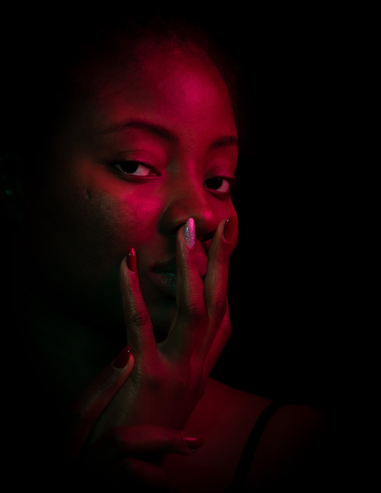 Portrait in red gel light. Both hands rest along her jaw, red nails against deep shadow, a level gaze.