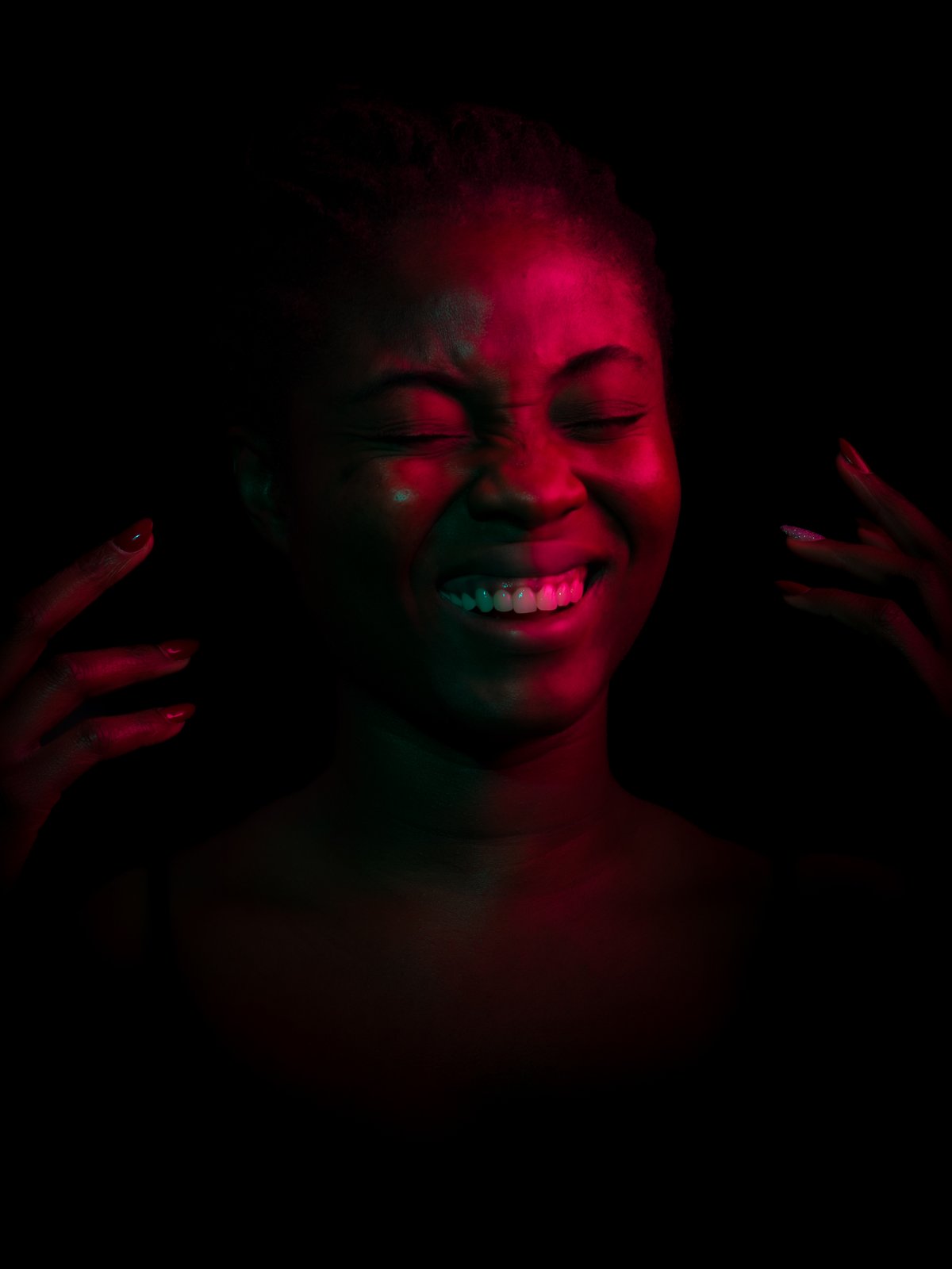 Portrait in red gel light. Eyes squeezed shut, a wide open laugh, hands raised beside her face.