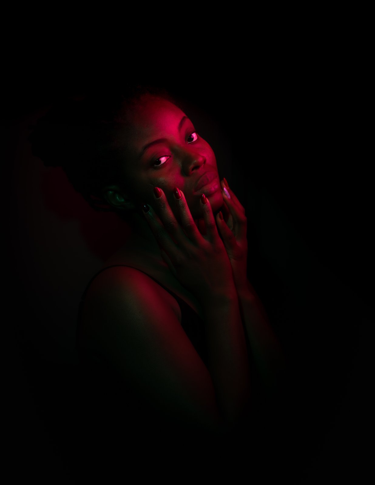 Portrait in red gel light with a green edge, hand raised to the mouth, a quiet look past the lens.