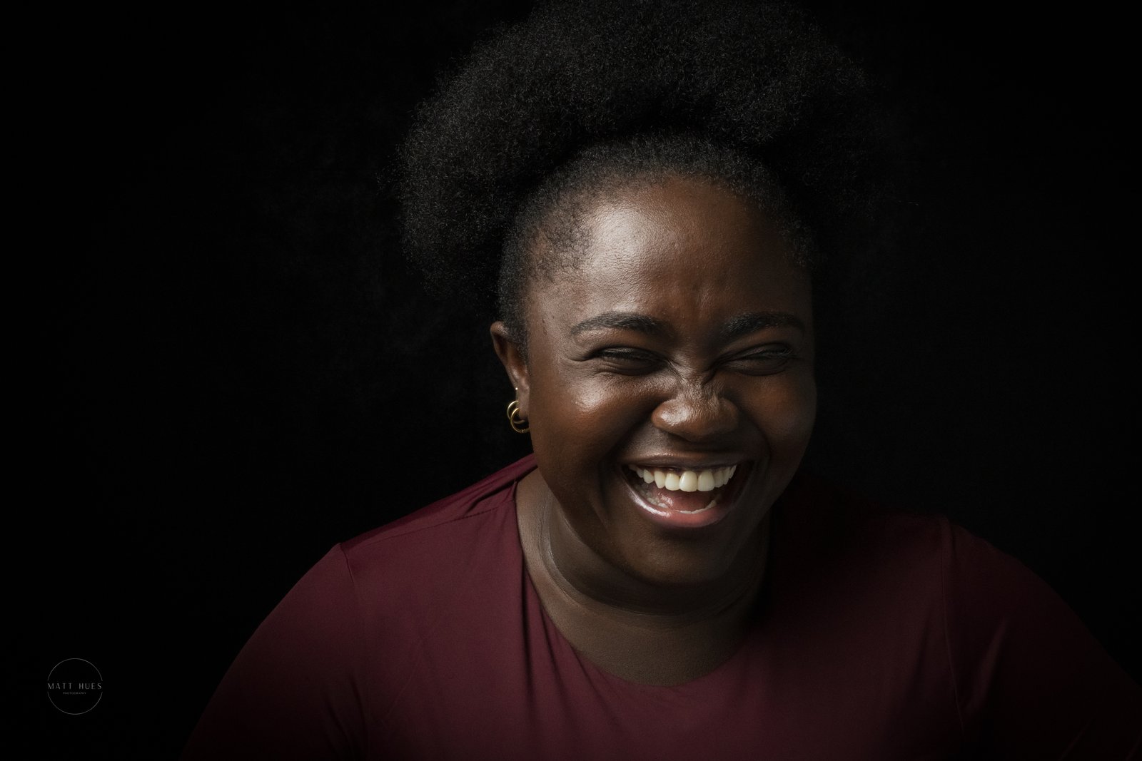 A woman laughs with her eyes squeezed shut against a dark studio backdrop, warm light on her face, hair pulled up.