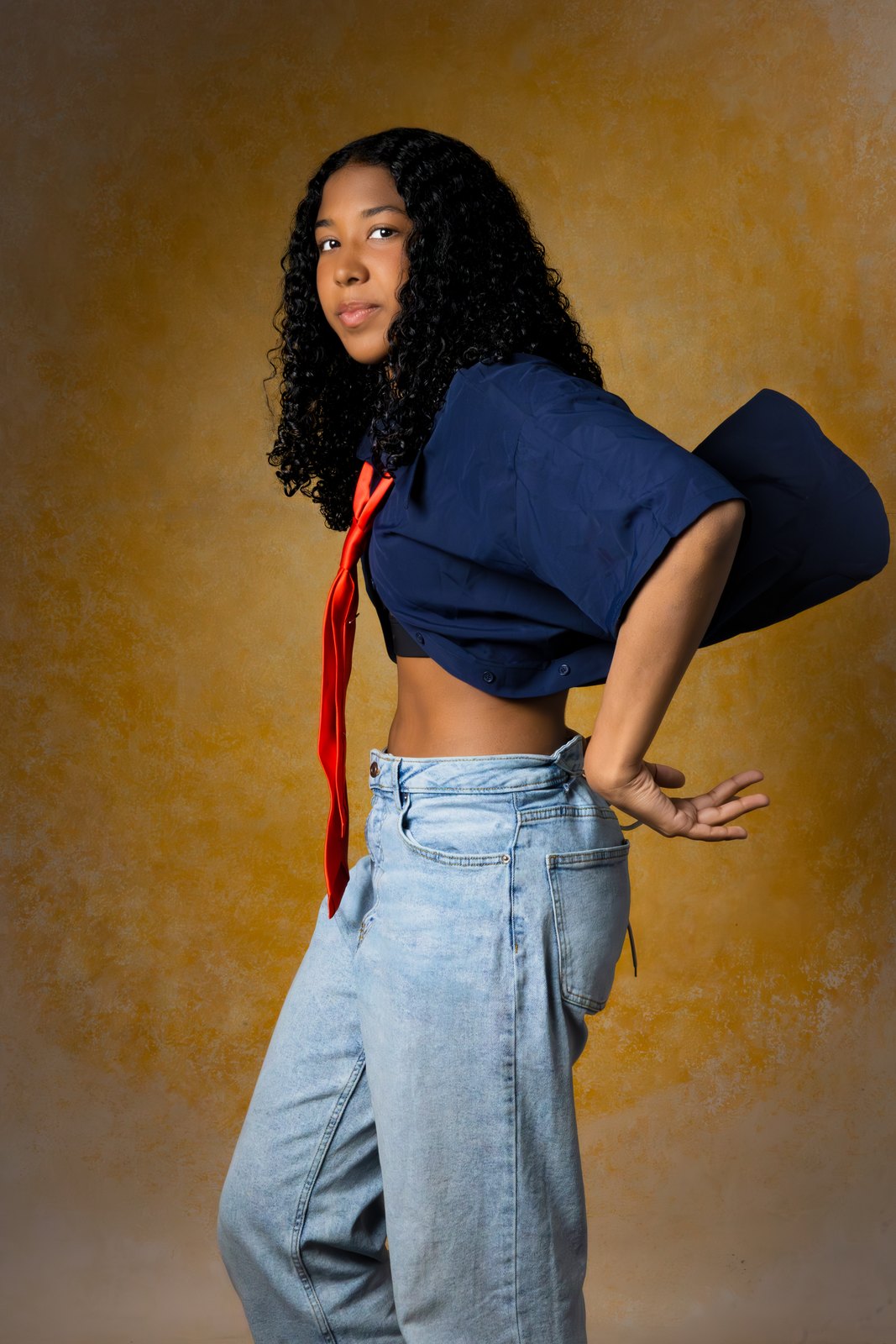 A young woman glances back in a cropped navy shirt and red satin tie on the Golden Sandstone backdrop.