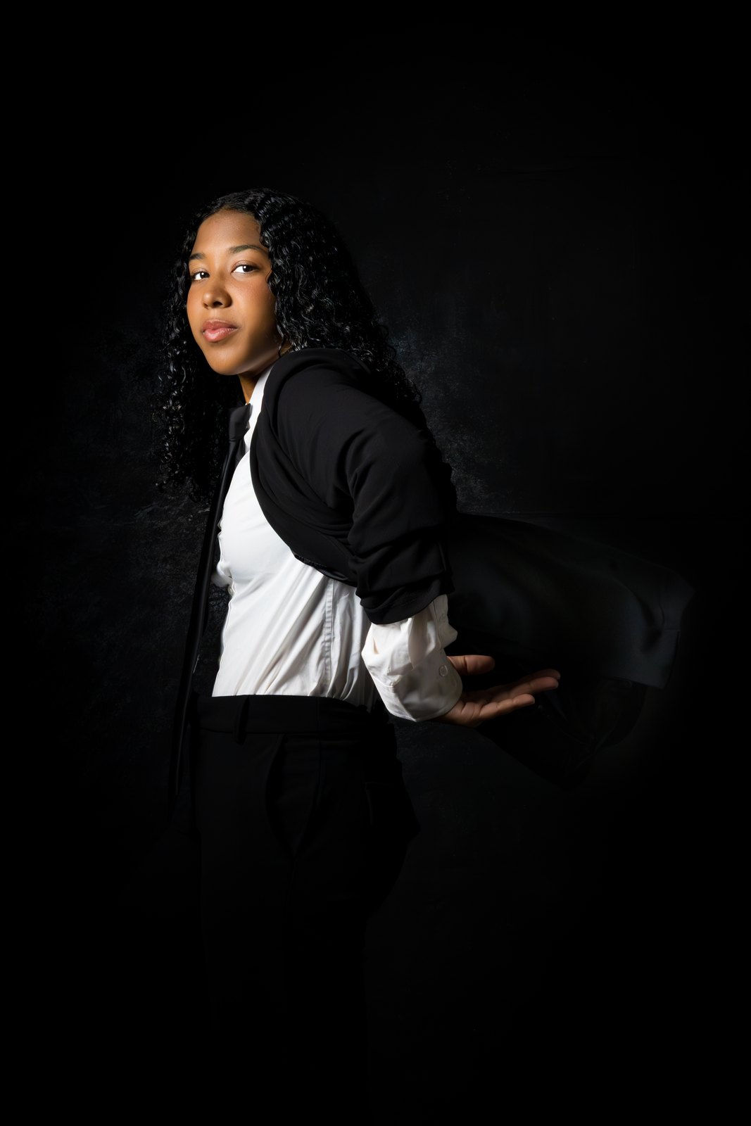A young woman in a black tux and white gloves, mid-gesture against deep charcoal.