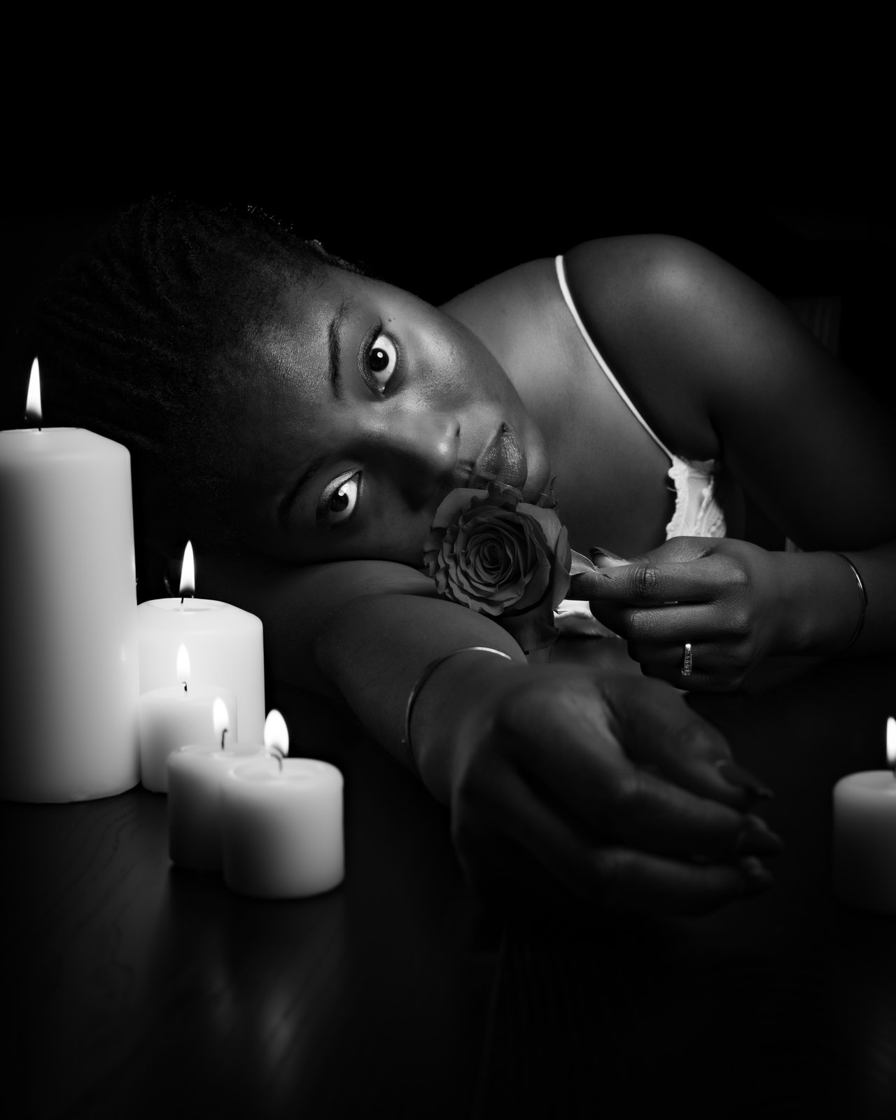 By candlelight, she rests her head on her arm with a red rose to her lips.