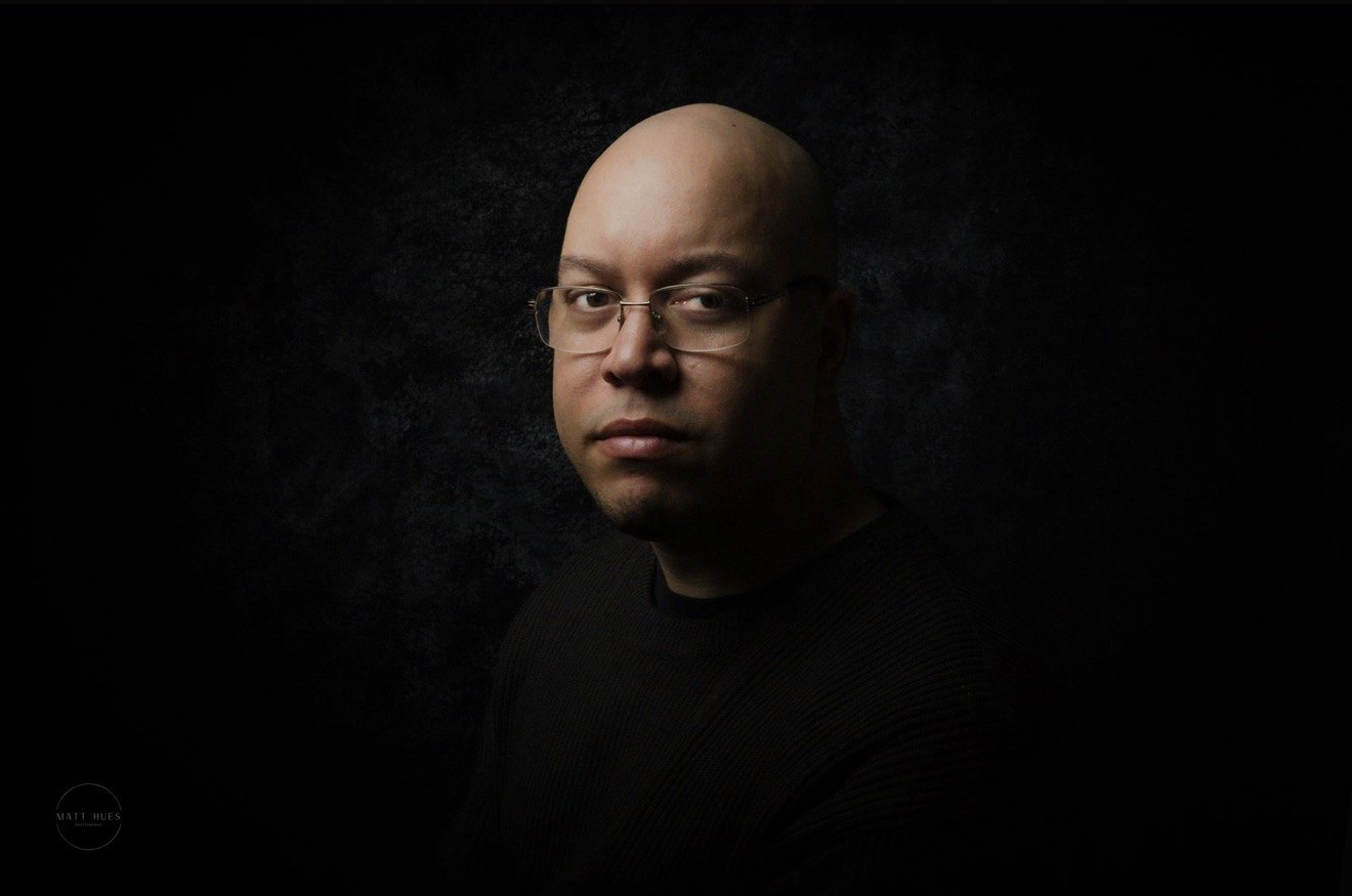 Matt Hues, side-lit in glasses and a dark sweater against a charcoal backdrop.