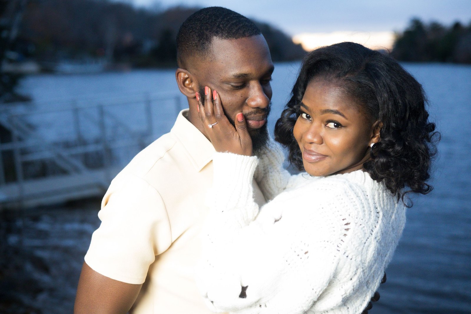 At dusk by the lake, she looks back at the camera, her hand with the ring resting on his cheek.