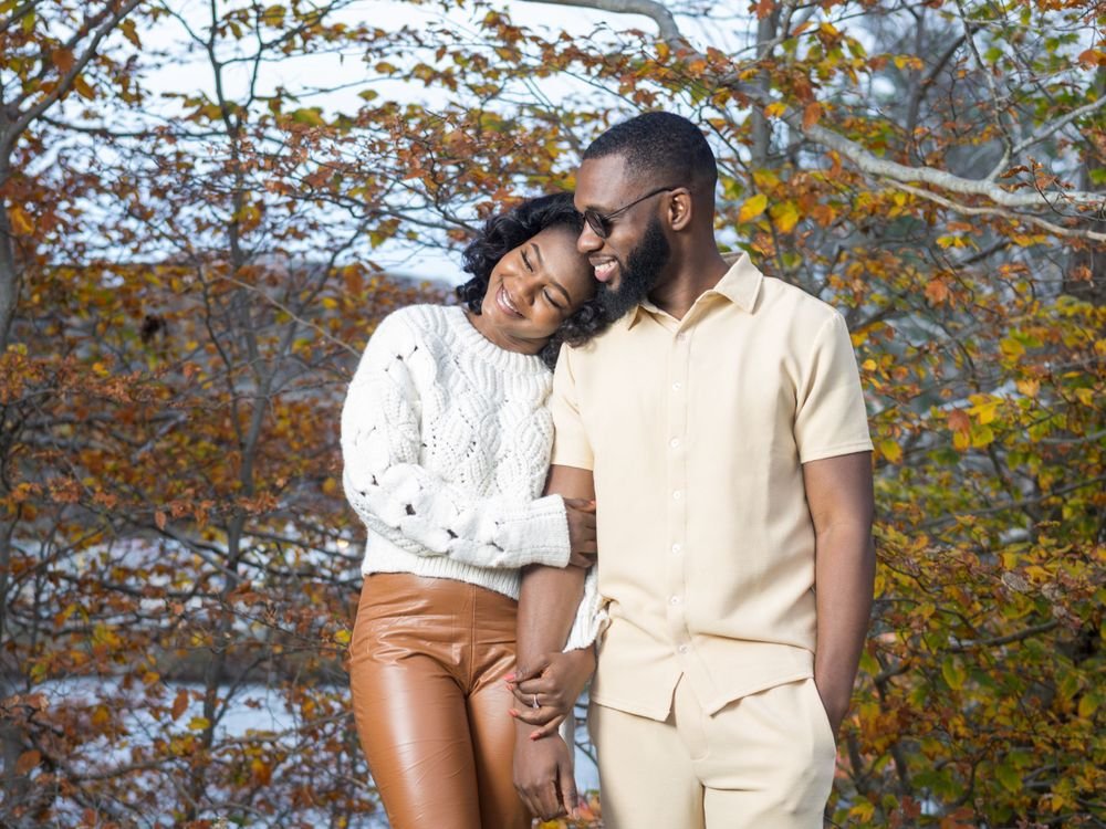 She leans into his shoulder with her eyes closed, smiling, as they hold hands under the autumn leaves.