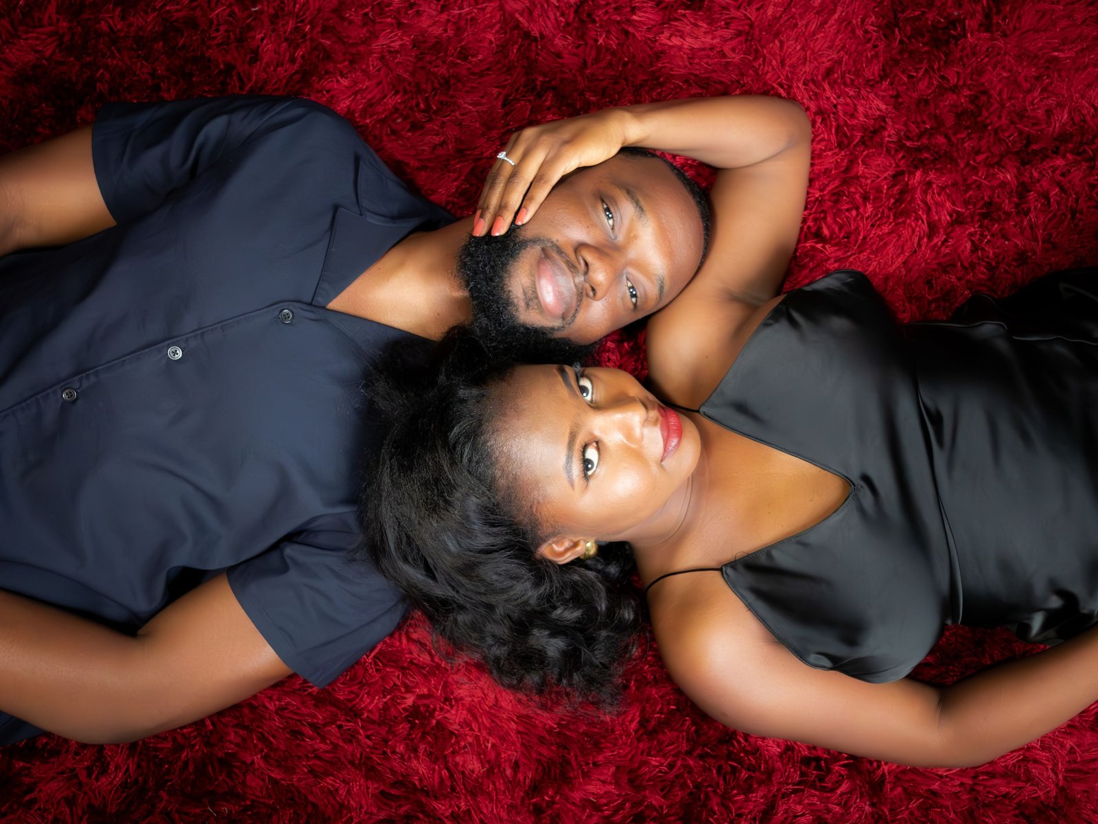 From above, the couple lies head to head on a deep red rug, her hand resting on his cheek.