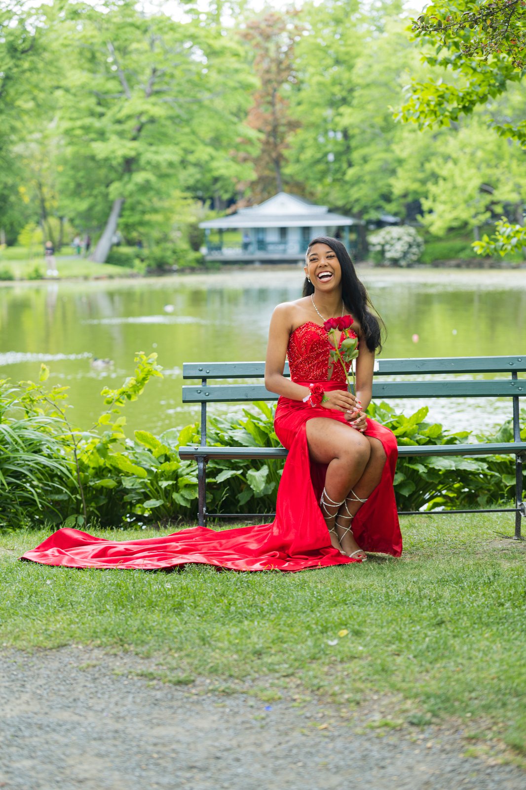 A graduate in a beaded red gown sits laughing on a park bench by the pond in the Halifax Public Gardens, her train spilling across the grass.