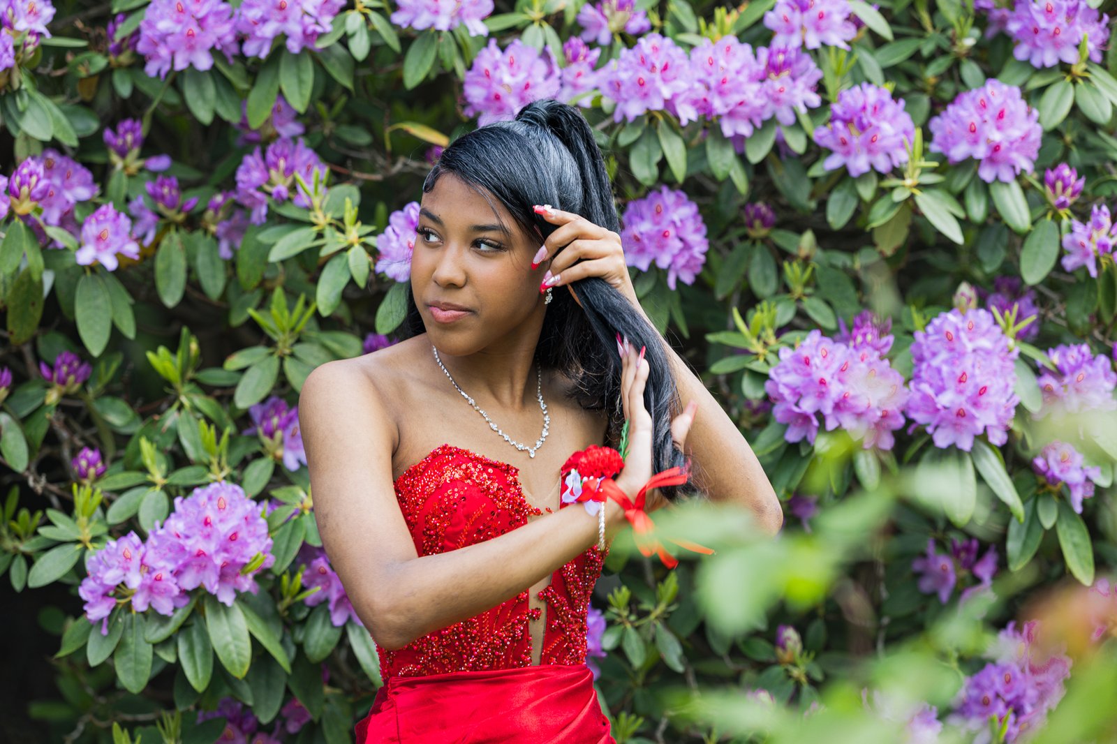 Close frame among the rhododendrons: she sweeps her hair back, red corsage tied at her wrist, looking away.
