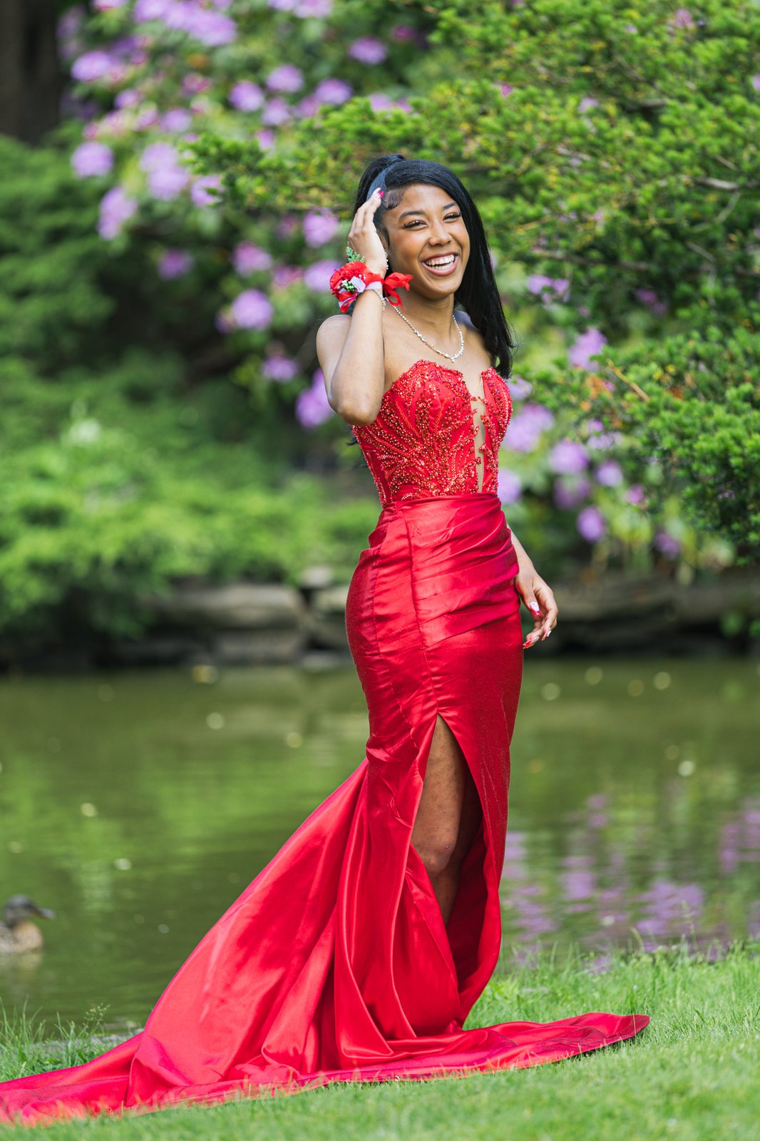 Standing by the pond in the red gown, mid-laugh, one hand raised to her hair, the train sweeping the grass behind her.