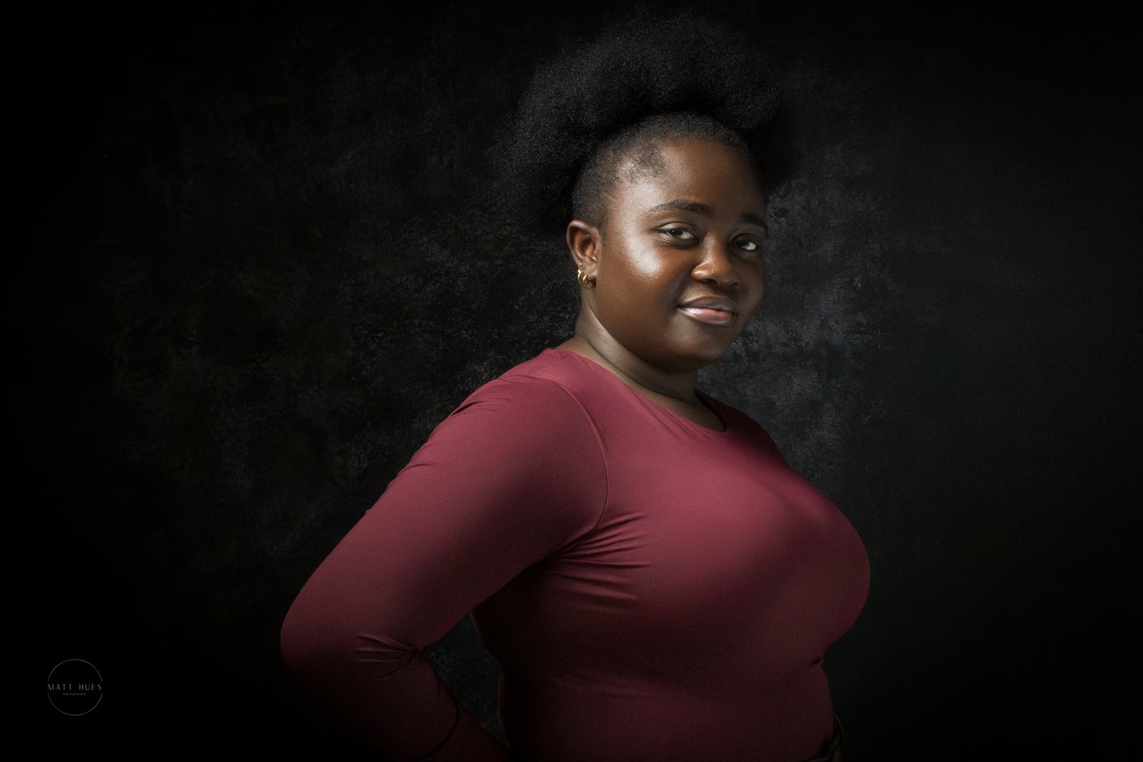 A woman in maroon turns with a hand on her hip, a knowing look on the dark backdrop.