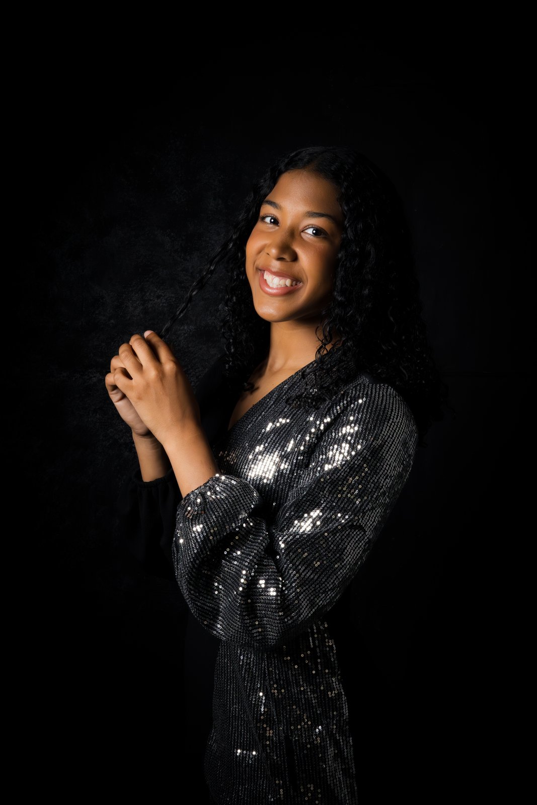 A young woman smiles in a sequined dress on charcoal, twirling a curl of hair.