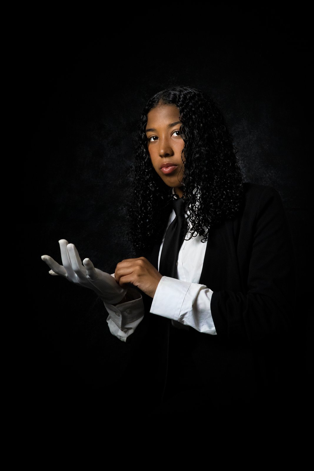 A young woman in black with a sequined sleeve, hands clasped, side-lit on charcoal.