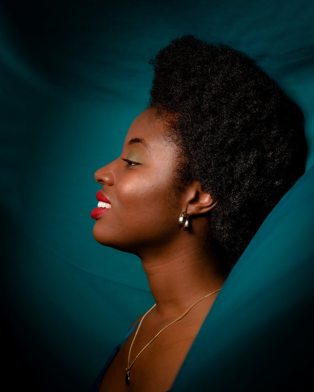 A profile against a sweep of teal fabric, red lip and gold hoop, eyes closed.