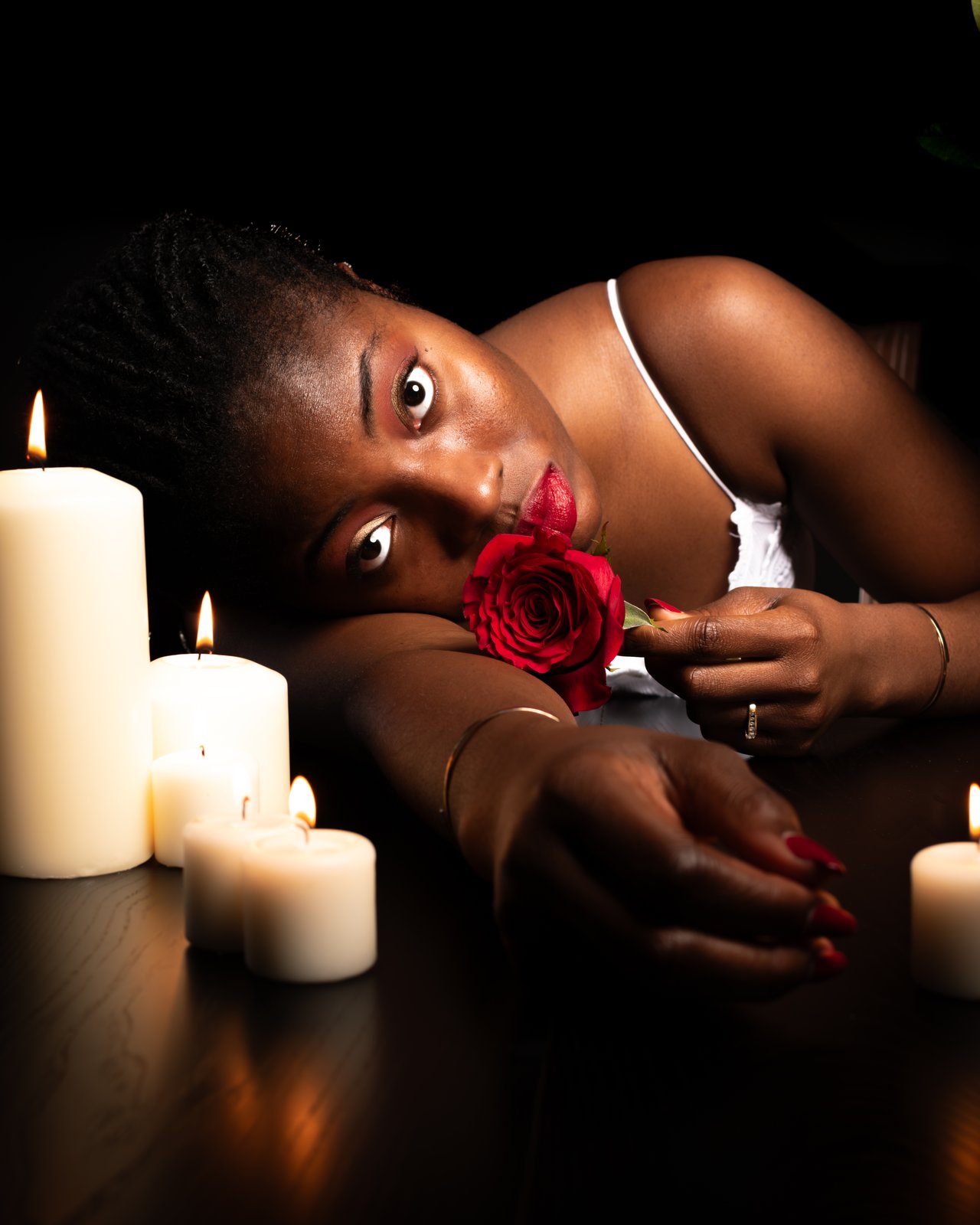 By candlelight, she rests her head on her arm with a red rose to her lips.
