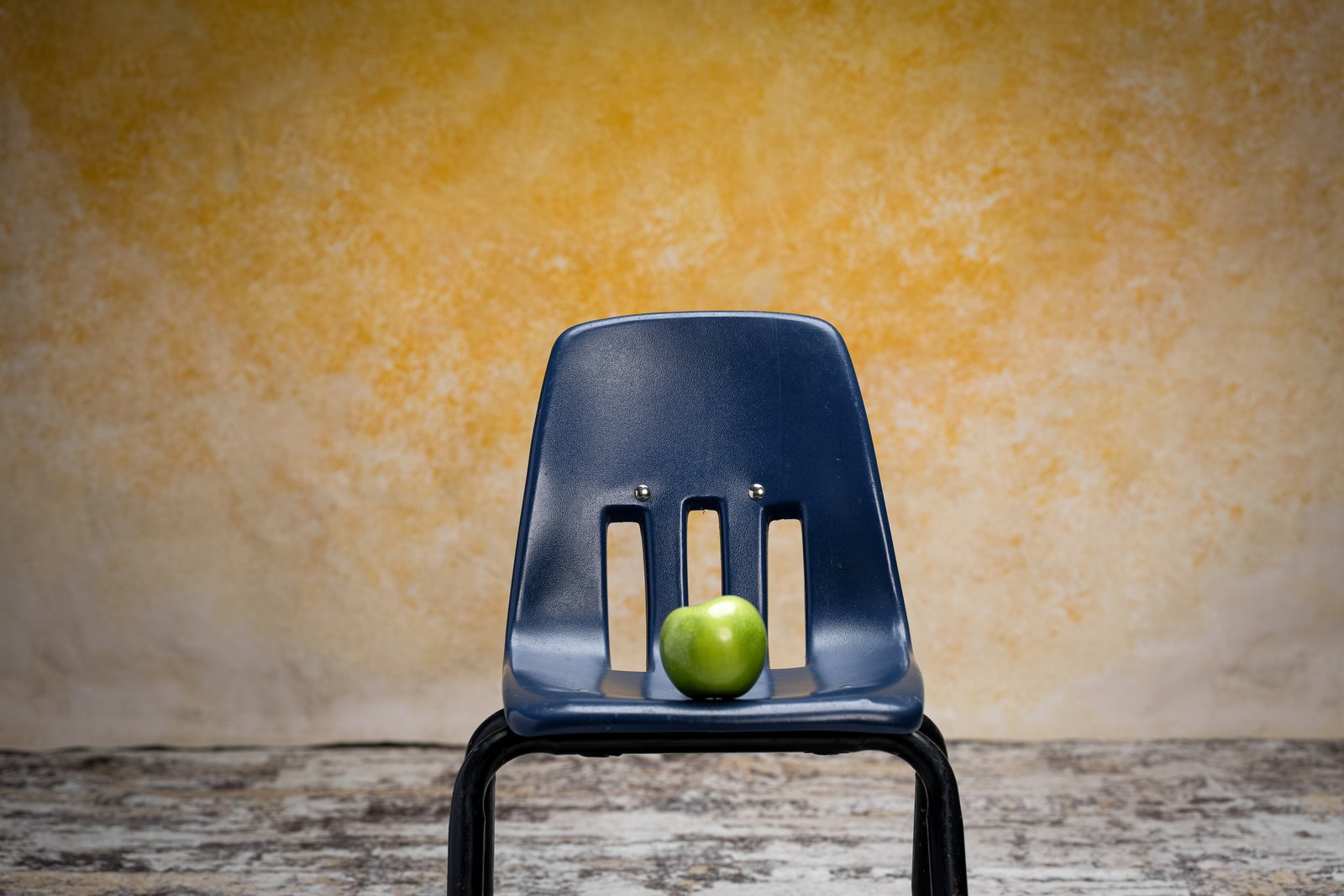 A green apple sits on a navy school chair in front of the hand-painted Golden Sandstone backdrop.